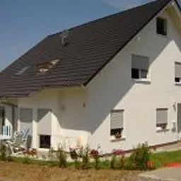 Biały, dwukondygnacyjny dom z ciemnym dachem, roletami w oknach i jasnymi elementami wykończeniowymi. Na pierwszym planie trawnik z młodymi roślinami, w tle widoczny fragment sąsiedniego budynku.